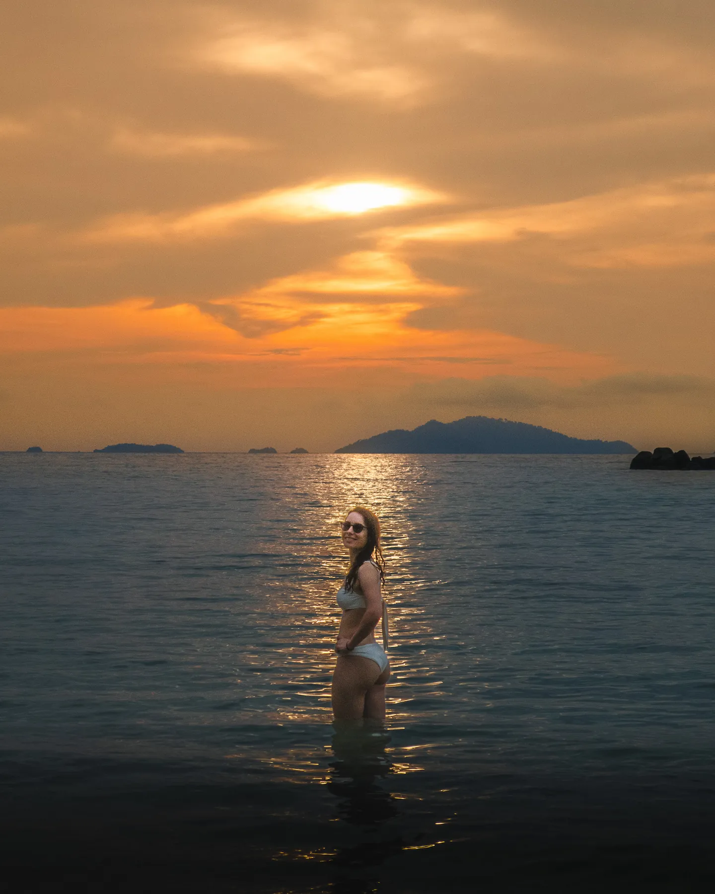 Woman standing in the water smiling at the camera with a sunset behind her