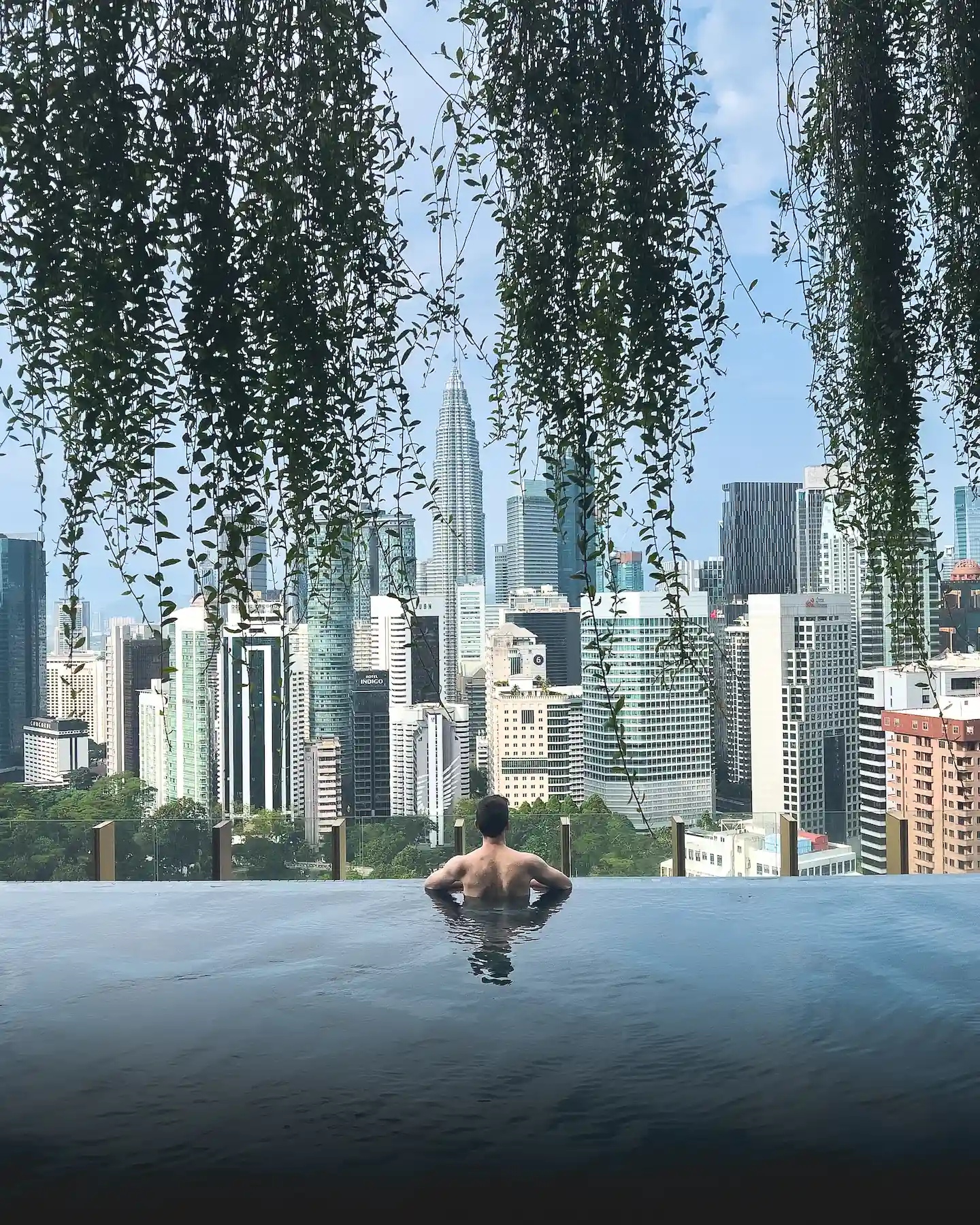 Back view of man in infinity pool with vines hanging from above and view of Kuala Lumpur skyline in background